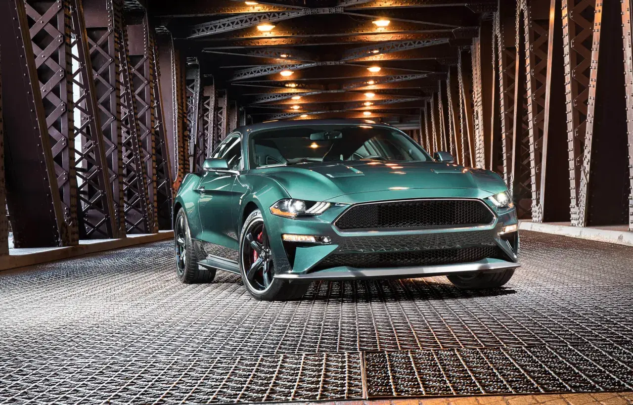 En grön pony car, en Ford Mustang, står på ett gallergolv under en bro med belysning.