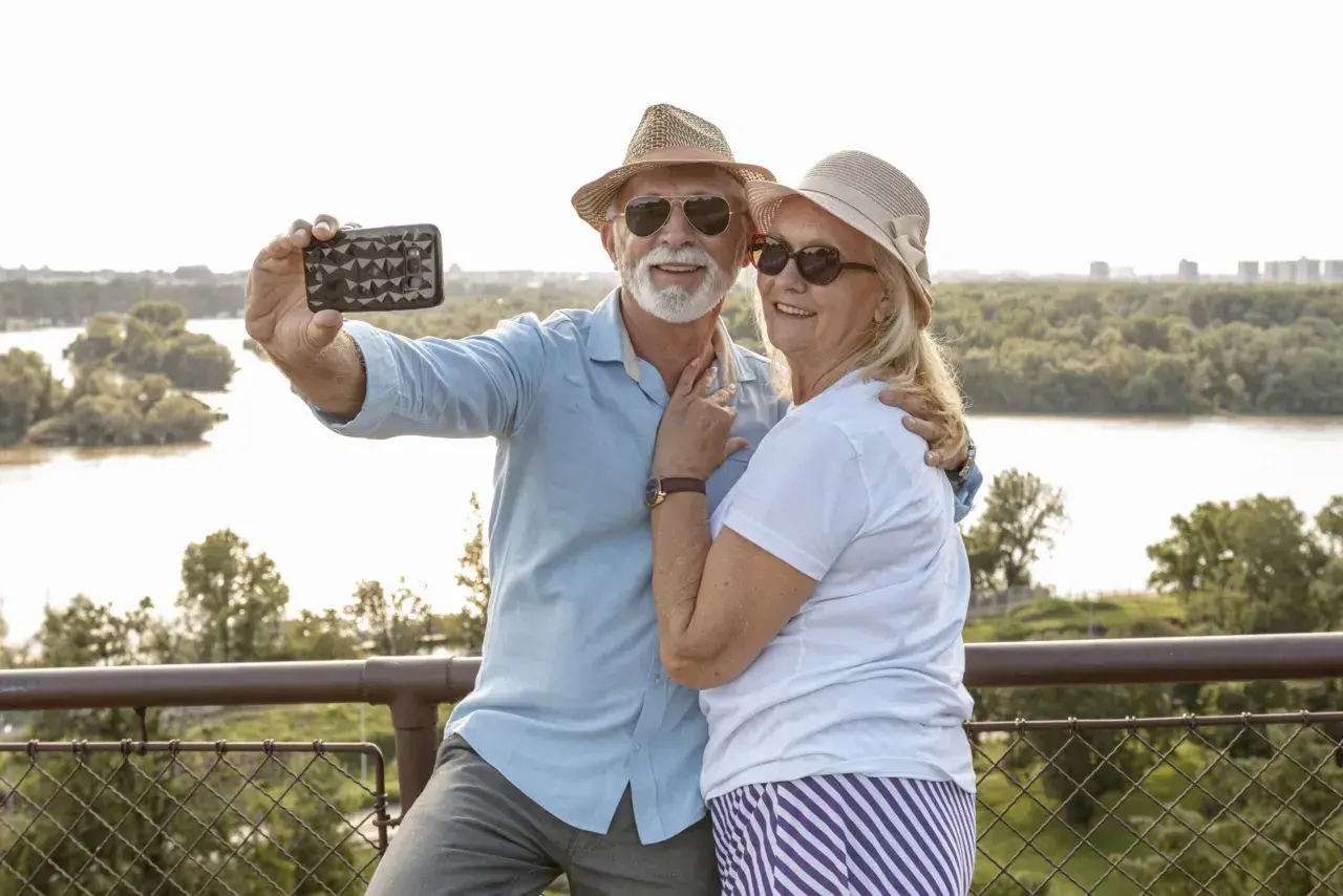 Uśmiechnięta para seniorów robi selfie na tle rzeki. Idealne wycieczki dla seniorów samolotem to świetny sposób na podróżowanie.