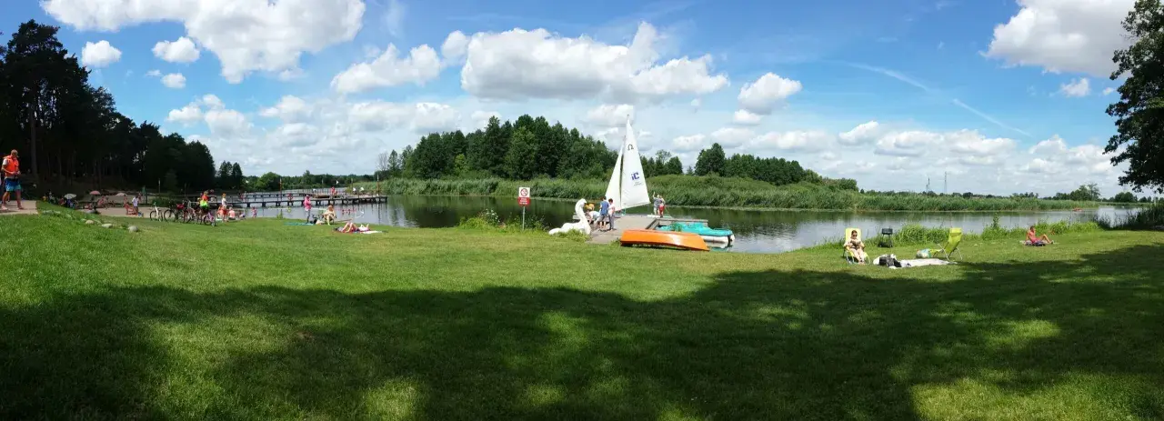 Słoneczny dzień nad jeziorem Kozienickim. Ludzie odpoczywają na trawie, opalają się, a na wodzie pływa żaglówka.