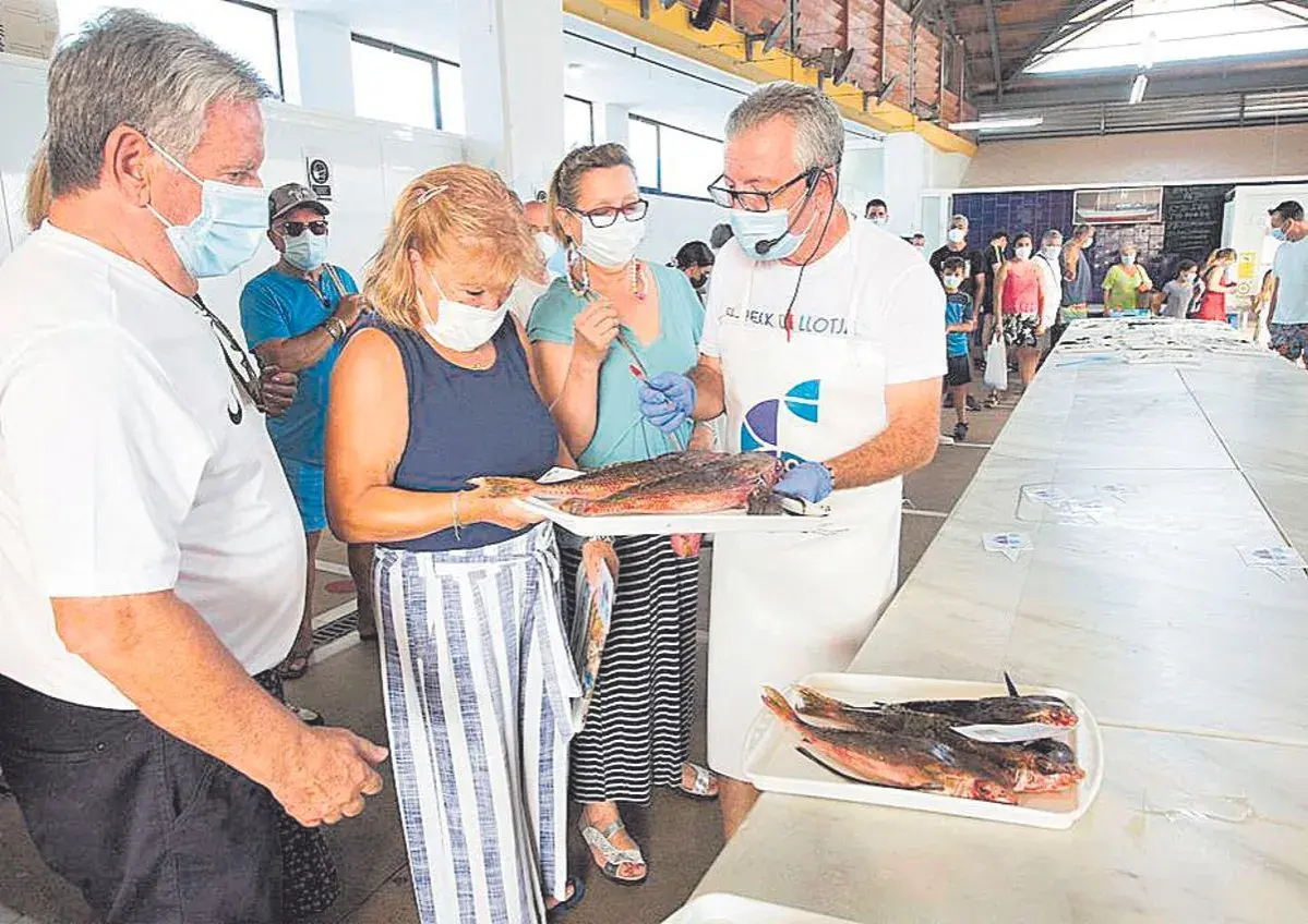 En la lonja del pescado, un hombre con delantal muestra unos peces a una mujer. Otras personas observan.