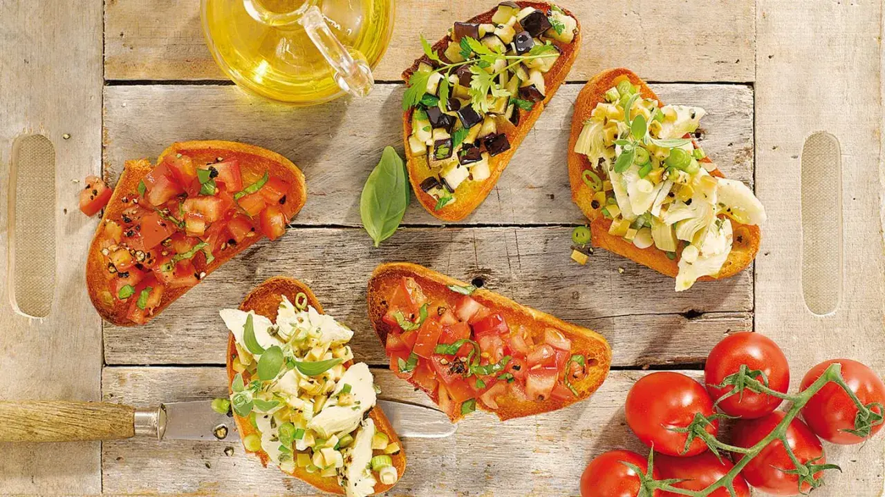 Fünf Bruschetta-Varianten mit Tomaten, Auberginen und Artischocken auf Holzhintergrund.