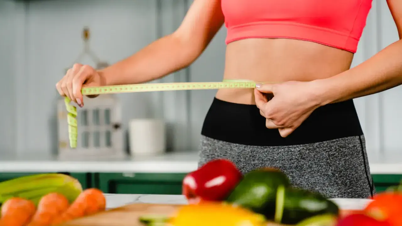 Mujer mide su cintura con cinta métrica, rodeada de verduras frescas. ¿La menopausia adelgaza o engorda?