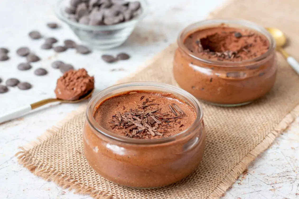 Deux pots de mousse au chocolat, l'un garni de copeaux de chocolat noir, sur une toile de jute. Une cuillère dorée présente une portion de cette délicieuse mousse.