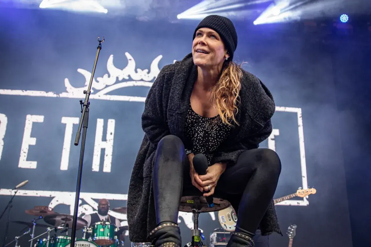 Beth Hart na scenie, w czapce i swetrze, śpiewa swoje utwory. W tle logo z koroną i perkusista.