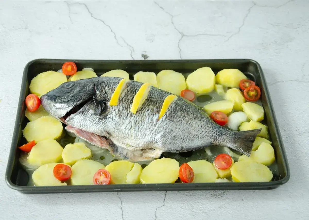 Un sargo pez entero, listo para hornear, rodeado de rodajas de patata y tomates cherry en una bandeja.