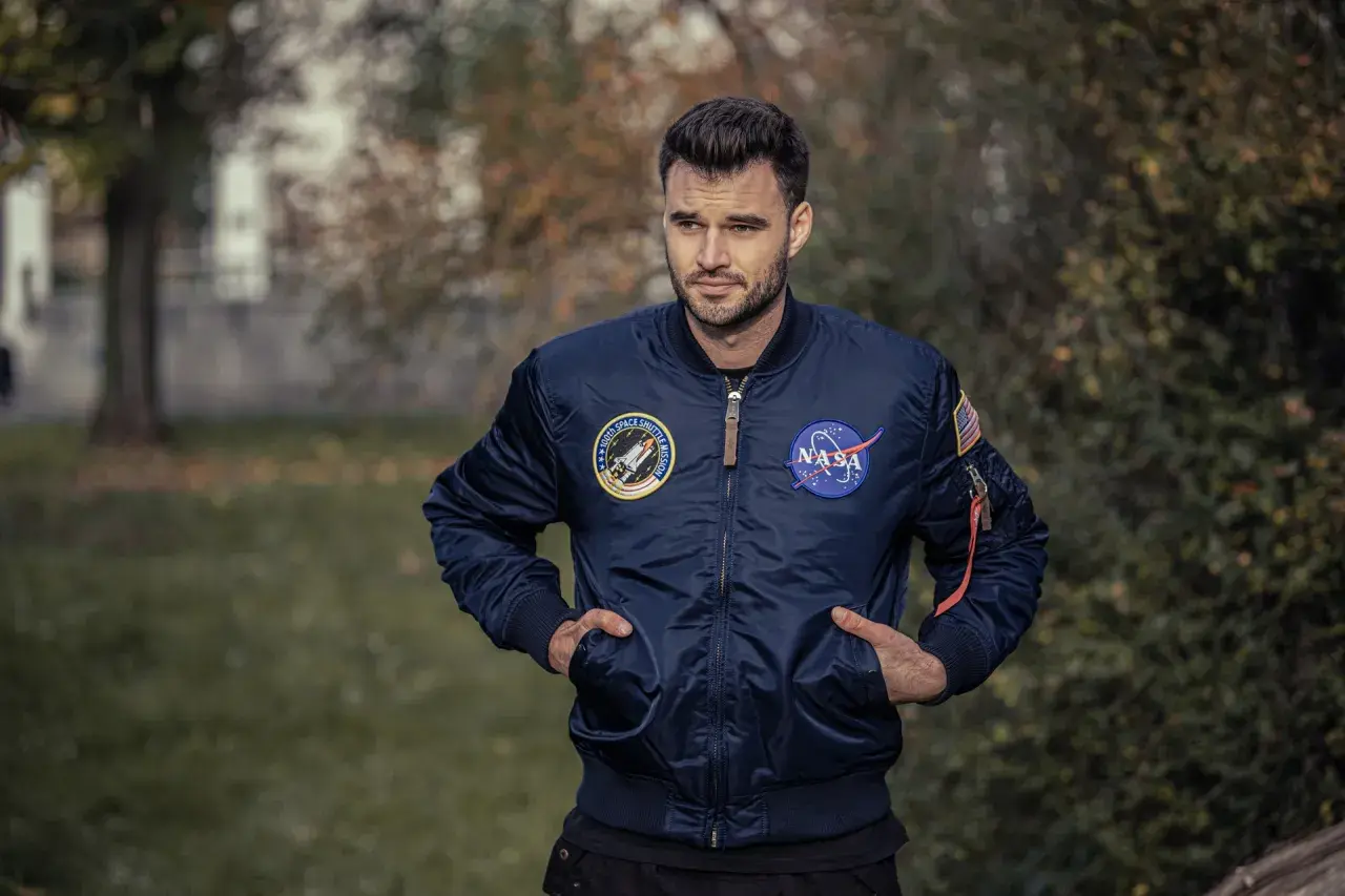 Mężczyzna w granatowej kurtce bomberce Alpha Industries z naszywkami NASA i misji wahadłowca. Alpha Industries to firma znana z odzieży inspirowanej lotnictwem.