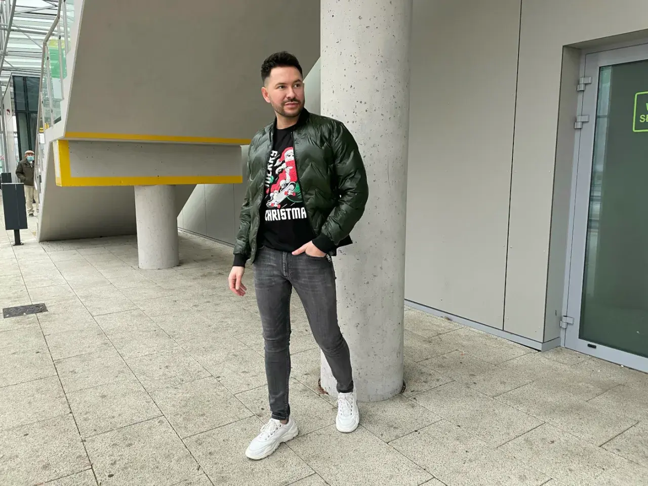 Mężczyzna w zielonej bomberce i szarych jeansach, zastanawia się, jakie buty do bomberki będą najlepsze. Postawił na białe sneakersy.