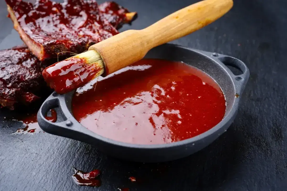 Pędzel z sosem barbecue zanurzony w miseczce, obok żeberka. Idealny sposób, jak zrobić sos barbecue.