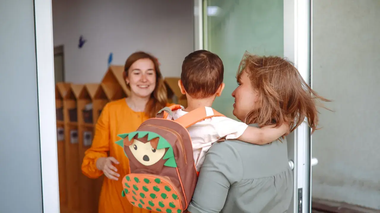 Madre abraza a su hijo con mochila de erizo, mientras una maestra sonríe. Inicio del periodo de adaptación infantil.
