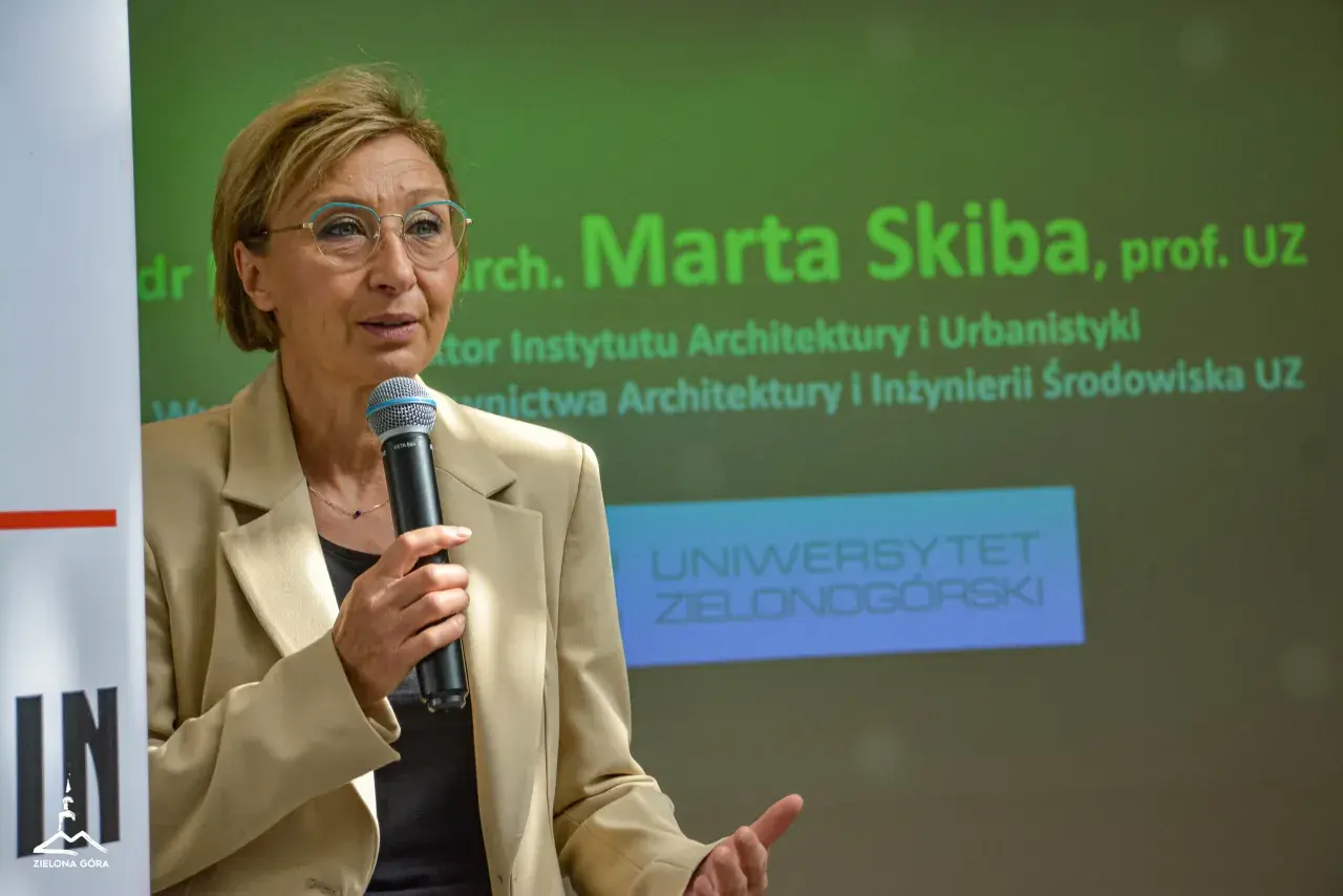 Kobieta w beżowej marynarce mówi do mikrofonu na tle zielonego ekranu z napisem "Uniwersytet Zielonogórski".