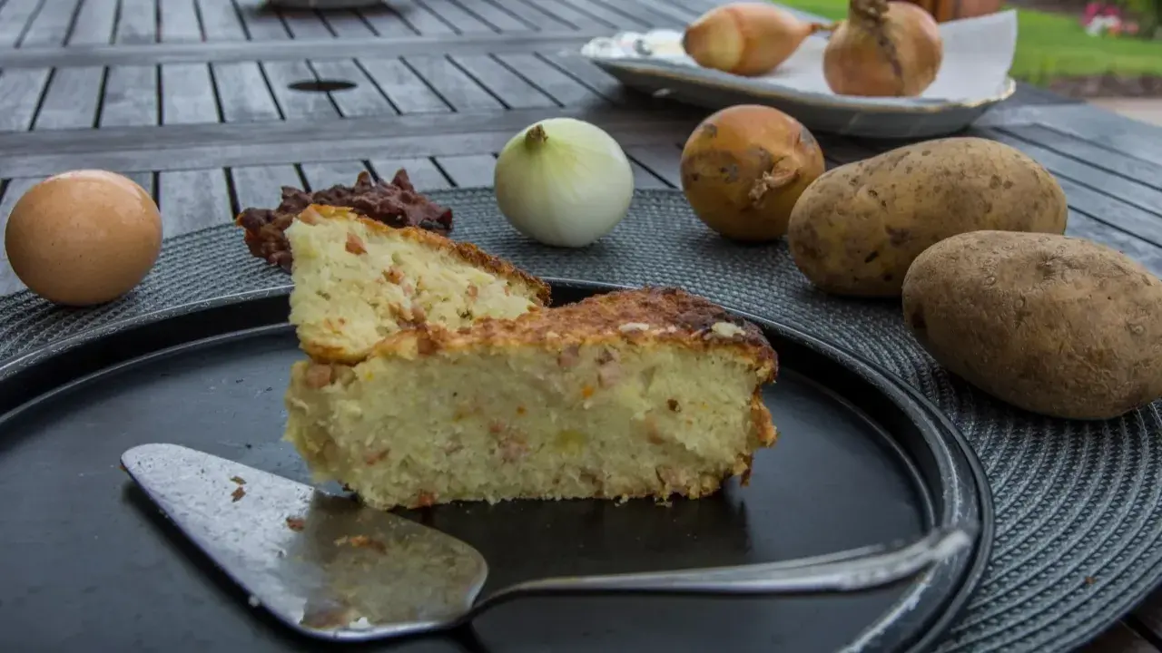 Babka ziemniaczana przepis babci: złocista, pachnąca babka ziemniaczana z boczkiem, obok surowe ziemniaki, cebula i jajko.