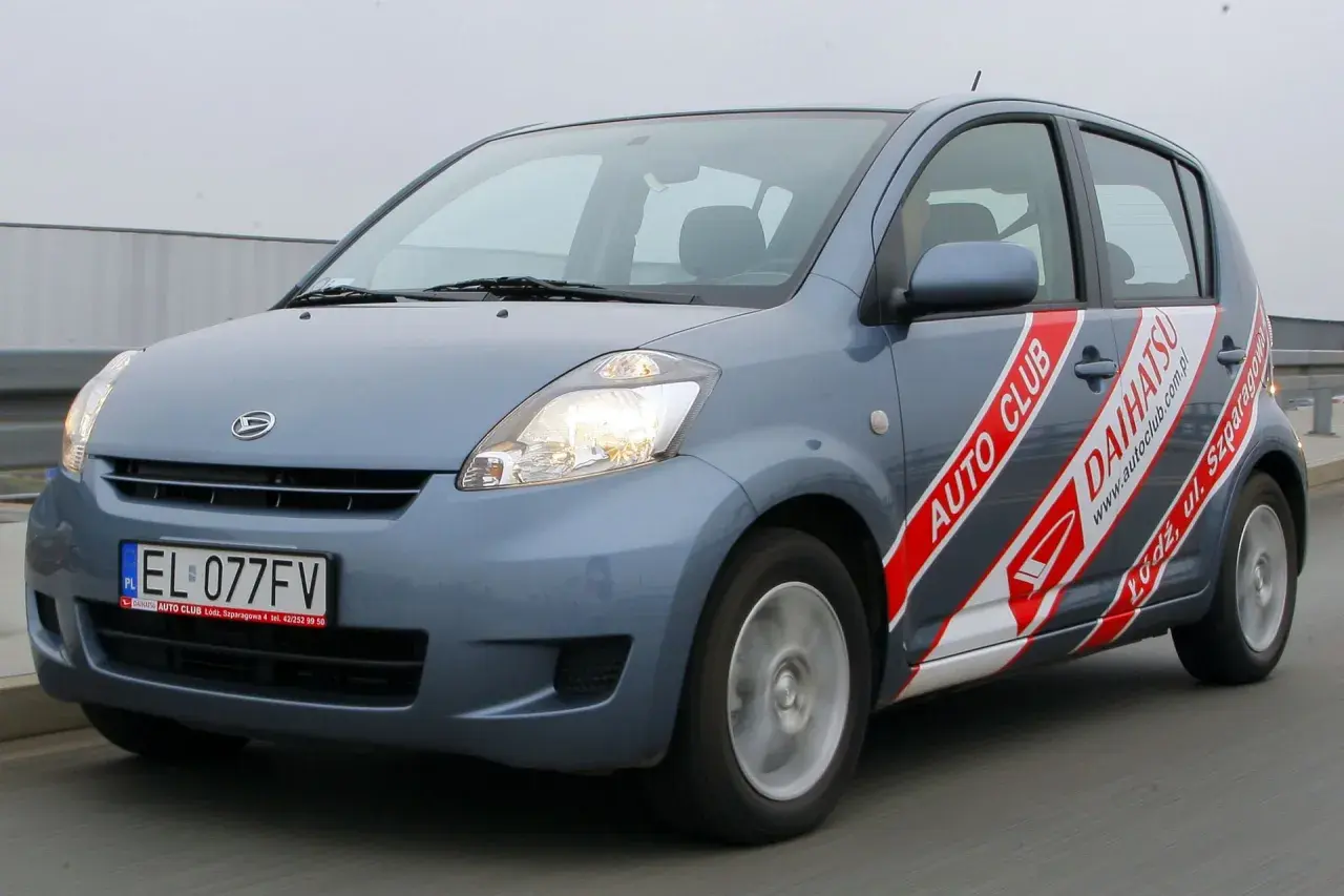 Szary Daihatsu Sirion z napisem "AUTO CLUB DAIHATSU" na boku, jadący po drodze.