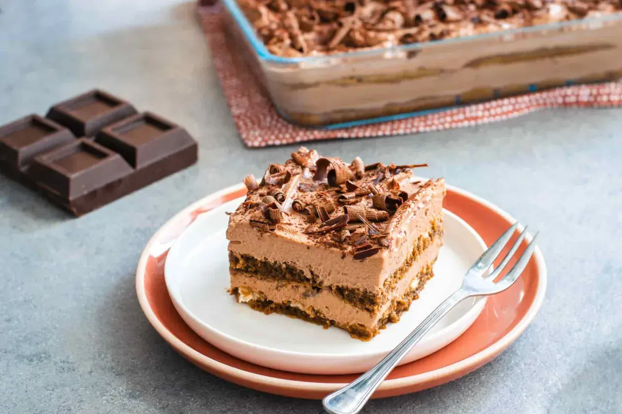 Una fetta di tiramisù cioccolato, ricca e cremosa, guarnita con riccioli di cioccolato. Accanto, pezzi di cioccolato fondente e un'intera teglia.