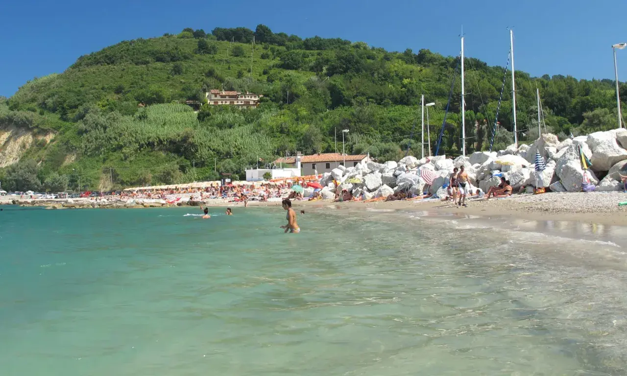 Spiaggia affollata con persone che nuotano e prendono il sole, sullo sfondo una collina verdeggiante. Gabicce Mare, dove si trova questo angolo di paradiso, è una perla dell'Adriatico.