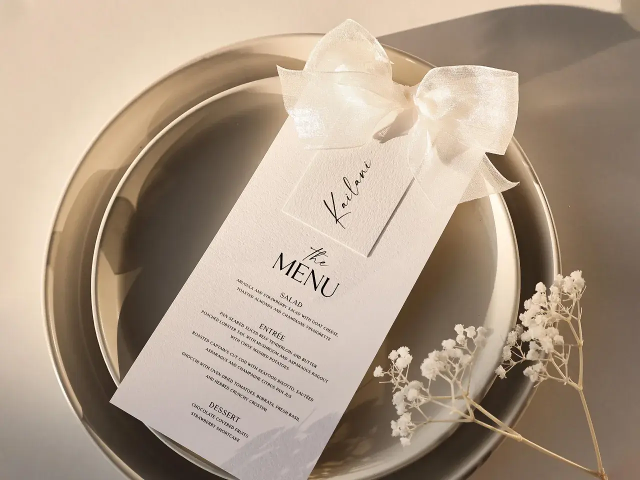 Menu de mariage élégant sur une assiette, orné d'un ruban et de petites fleurs blanches.