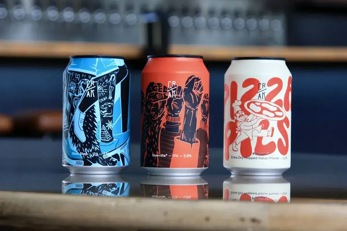 Tre lattine di crak beer con grafiche artistiche: una blu con piramide, una arancione con pugni alzati, una bianca con un pizzaiolo.
