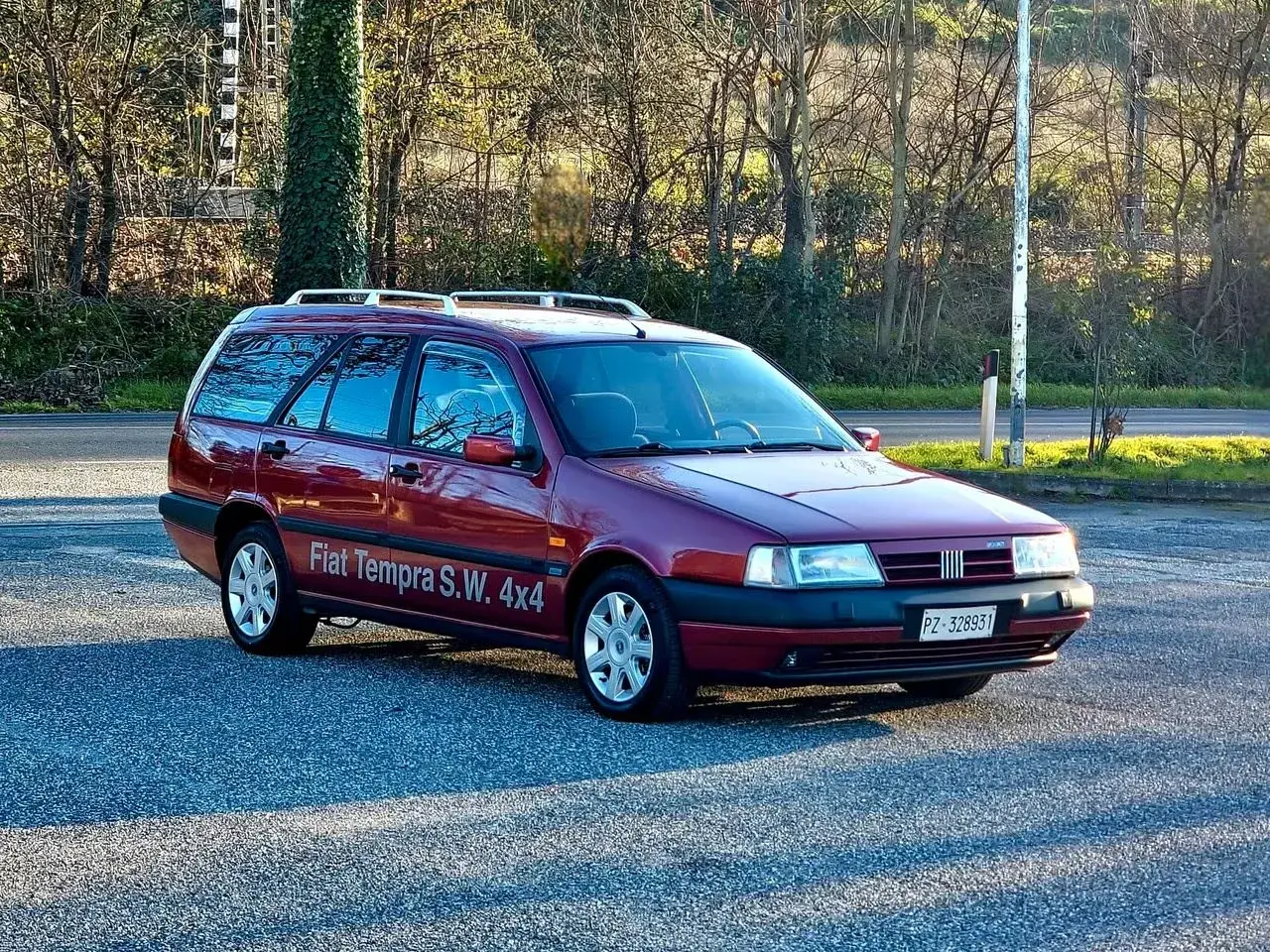 Ciemnoczerwony Fiat Tempra S.W. 4x4 z relingami dachowymi zaparkowany na tle drzew.