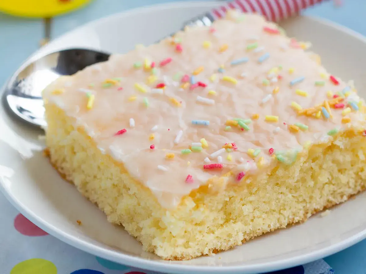 Ein Stück saftiger Fantakuchen vom Blech, mit Zuckerguss und bunten Streuseln bestreut, auf einem Teller mit Löffel.