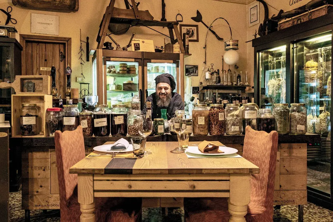 Un cuoco sorridente in un locale con salumi appesi e barattoli di conserve. Un'atmosfera unica per ristoranti particolari a Brescia e provincia.
