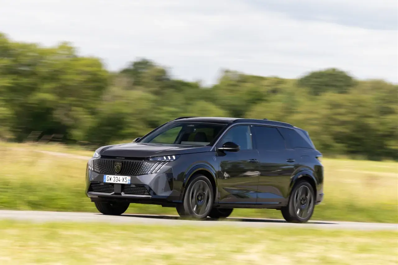 Ciemnoszary Peugeot 5008 pędzi po drodze, otoczony zielenią pól i drzew.