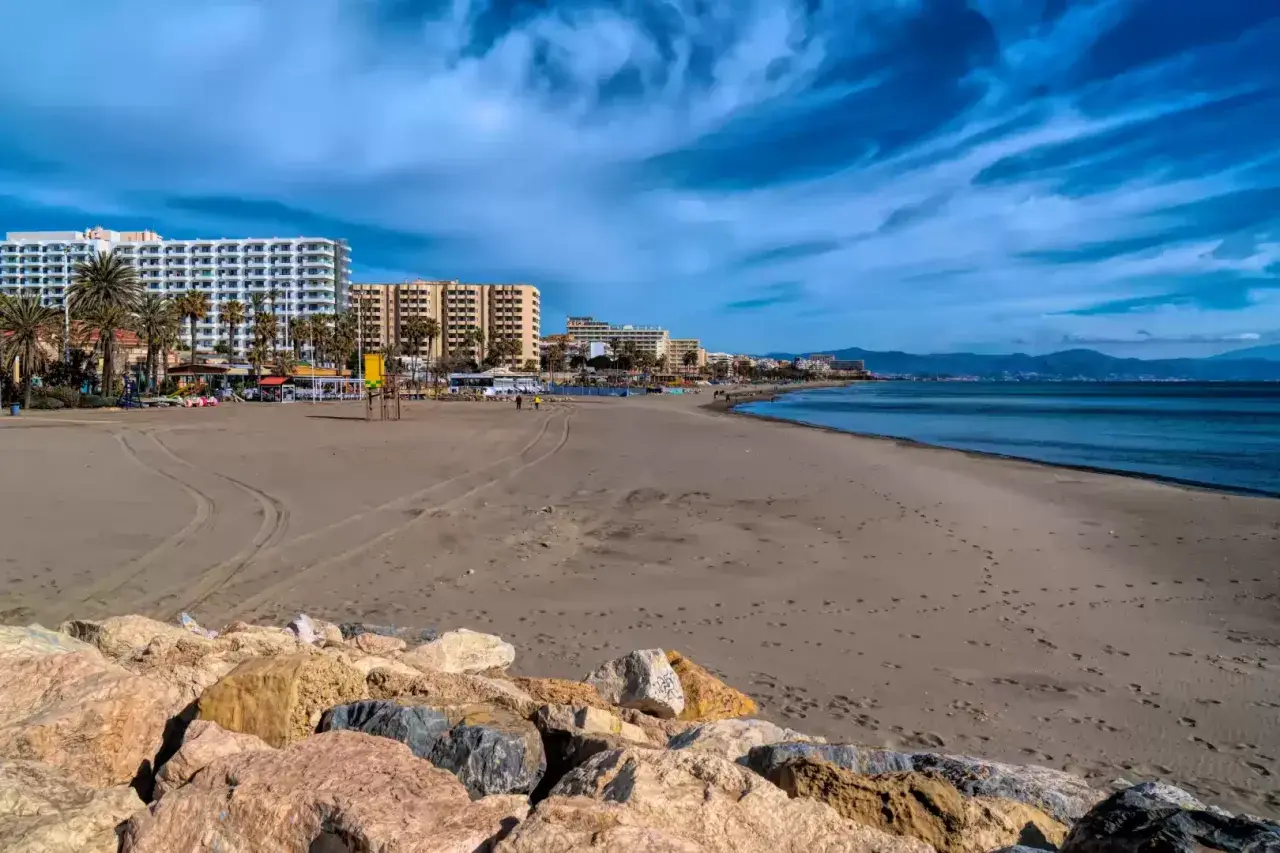 Amplia playa de arena con edificios hoteleros al fondo. El mar azul y el cielo nublado crean un paisaje sereno, ideal para pasear y sentir la **playa fuente de la salud**.