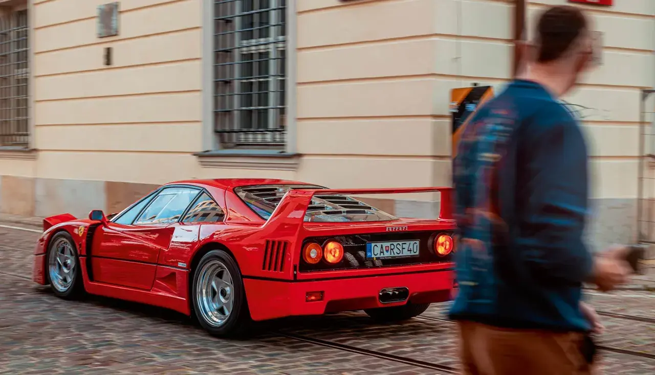 Czerwone Ferrari F40 z charakterystycznym spojlerem i okrągłymi światłami mija brukowaną ulicę, a obok przechodzi rozmyta postać mężczyzny.
