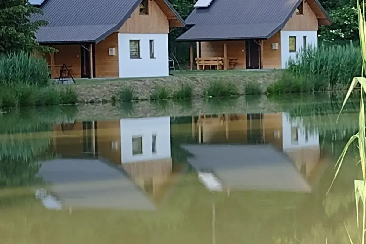 Urokliwe łowisko z domkami w Podkarpackiem. Dwa drewniano-białe domki z dachami odbijają się w spokojnej tafli wody.