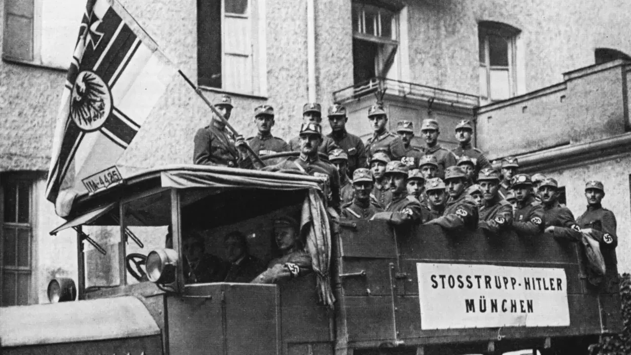 Männer in Uniformen auf einem LKW mit der Aufschrift "STOSSTRUPP HITLER MÜNCHEN". Ein Mann hält eine Fahne mit einem Adler. Dies ist ein Bild vom Hitlerputsch.