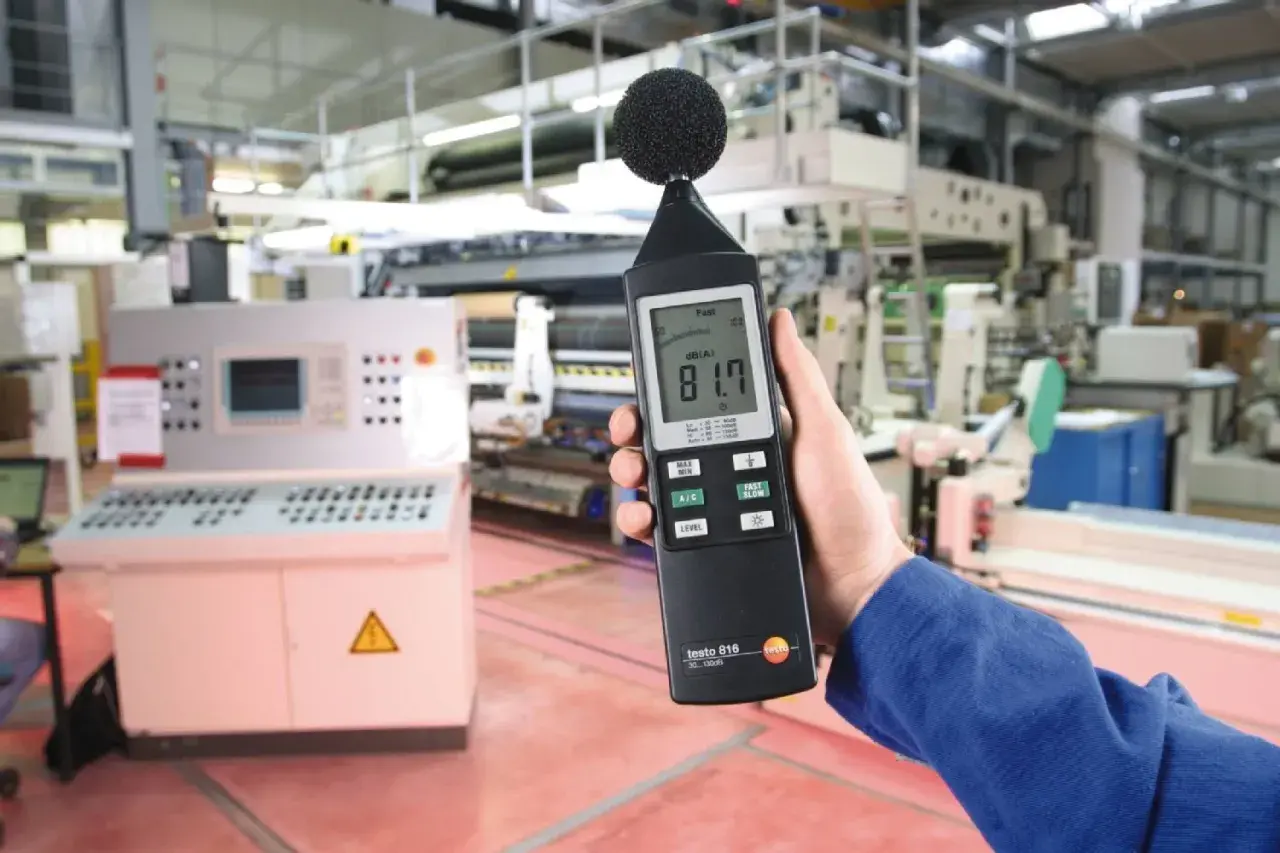 Mano sostiene sonómetro que mide 81.7 dB(A) en una fábrica, indicando el nivel de ruido.