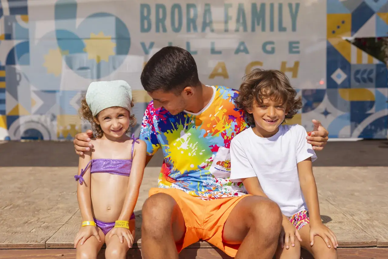 Un uomo con due bambini sorridono davanti a uno sfondo con la scritta "Brora Family Village".