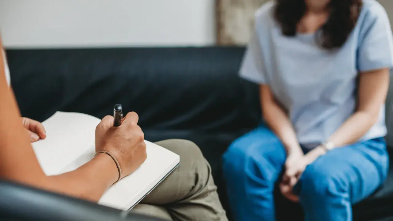 Eine Person schreibt in ein Notizbuch, während eine andere Person in blauen Jeans auf einer Couch sitzt. Dies könnte eine Szene der MDMA-Therapie sein, die bei der Heilung hilft.
