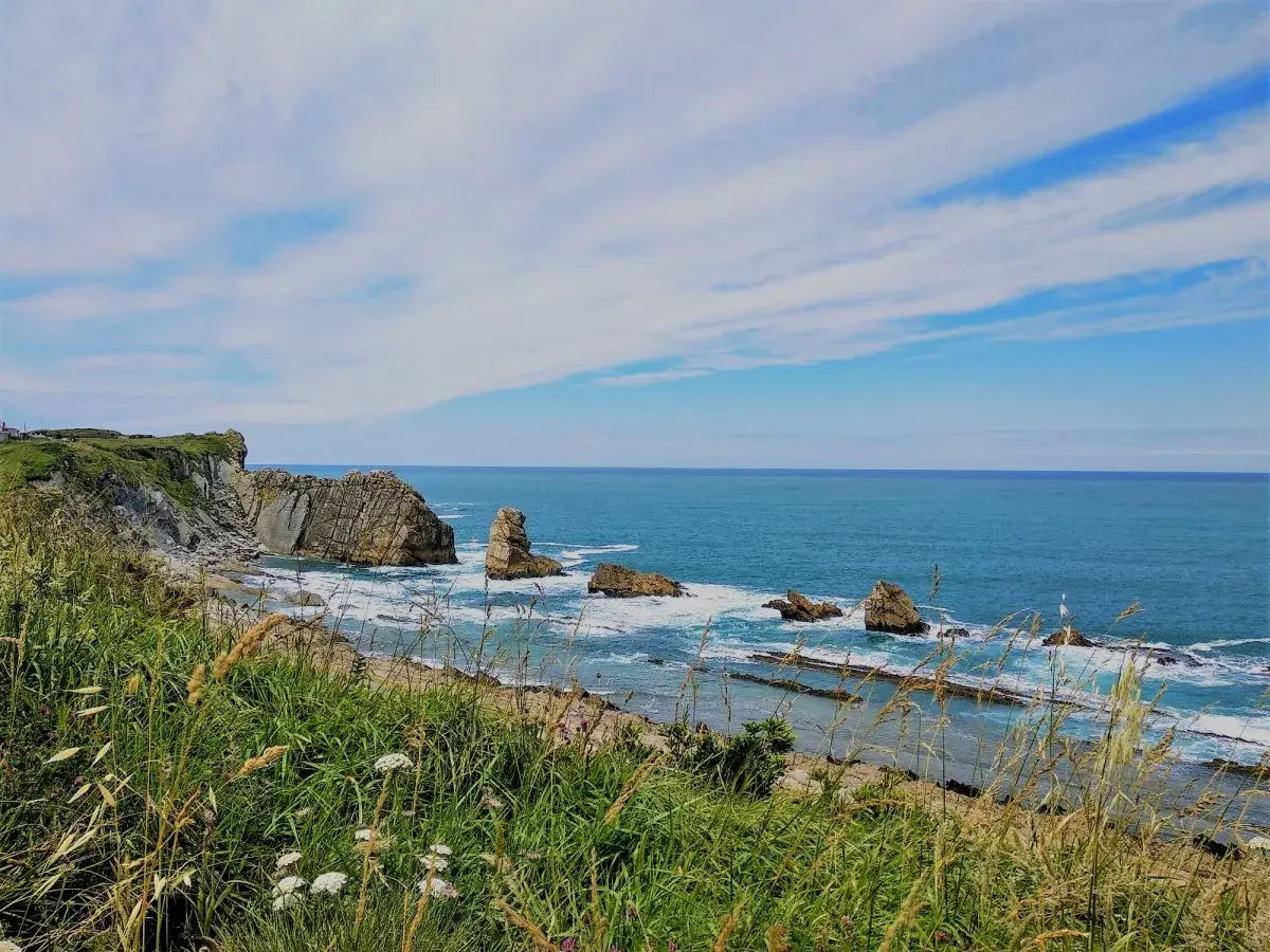 Vista de la **playa de dicido** con rocas emergiendo del mar azul y vegetación verde en primer plano bajo un cielo nublado.