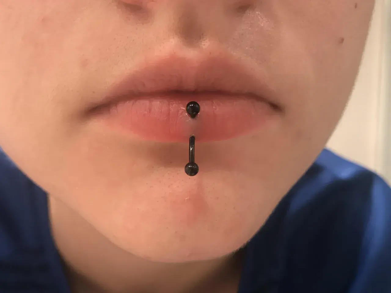 Osoba z pionowym kolczykiem w wardze (vertical labret piercing). Czarny, błyszczący kolczyk przechodzi przez środek dolnej wargi.