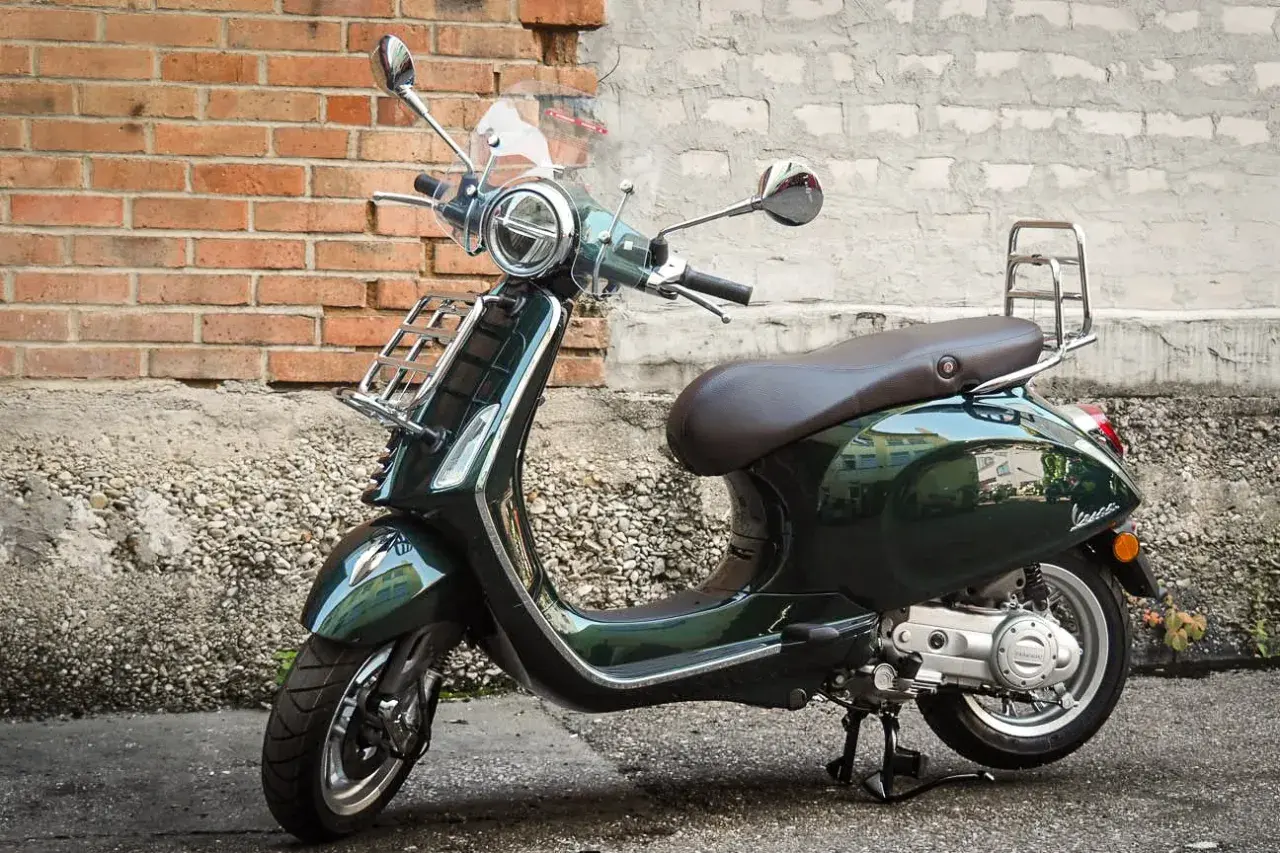 Elegancki, ciemnozielony skuter Piaggio z brązowym siedzeniem i chromowanymi dodatkami stoi na tle ceglanej ściany.