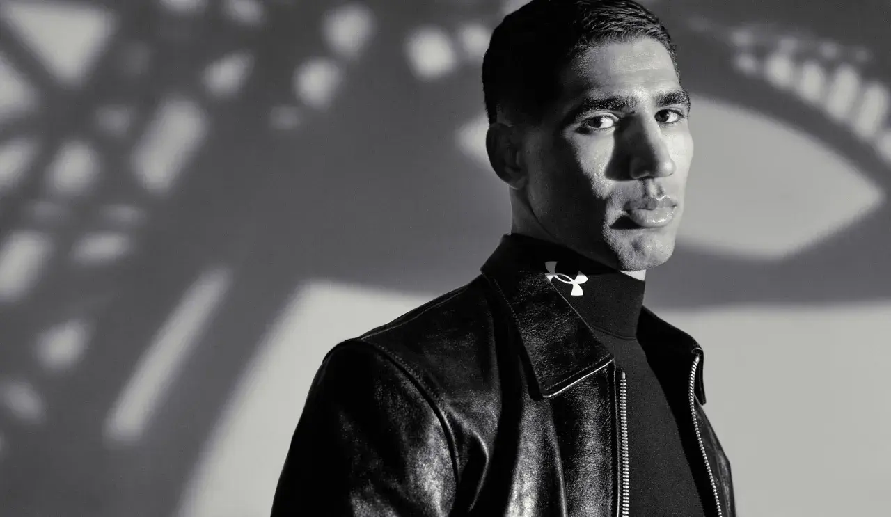 Achraf Hakimi, ein Fußballstar, blickt nachdenklich in die Kamera, trägt eine Lederjacke und ein schwarzes Rollkragenshirt mit dem Under Armour Logo.