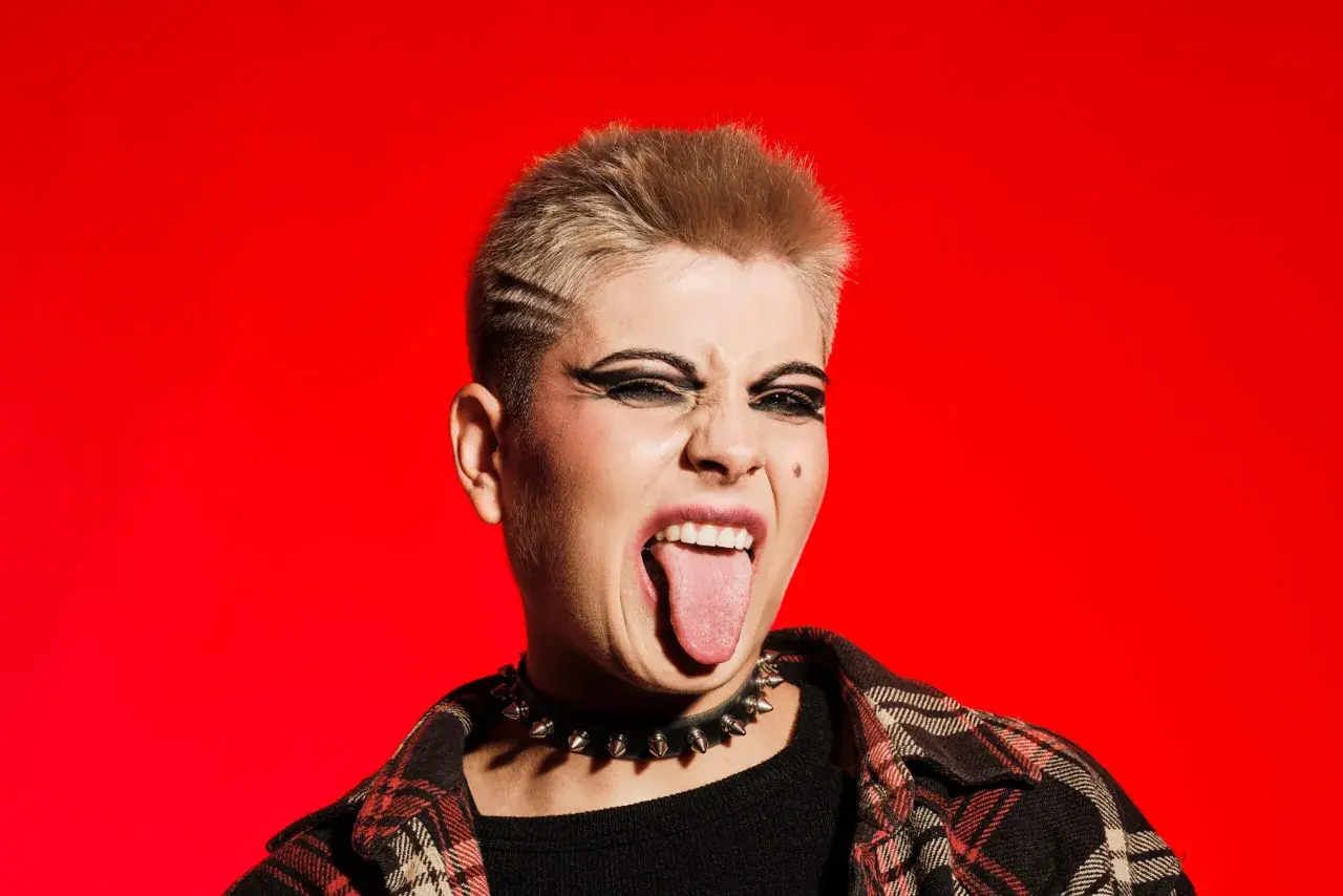 Młoda osoba z blond włosami i mocnym makijażem oczu, w kolczatce i flanelowej koszuli, wyciąga język. To kwintesencja buntu i stylu punk.