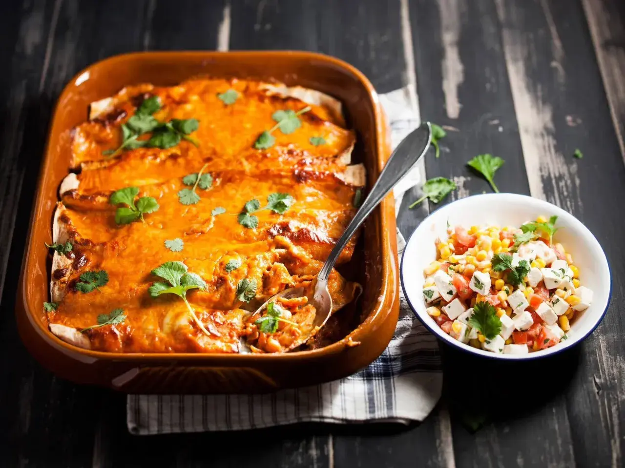 Enchiladas kyckling i en ugnsform, toppade med ost och koriander, bredvid en skål med majs- och tomatsallad.