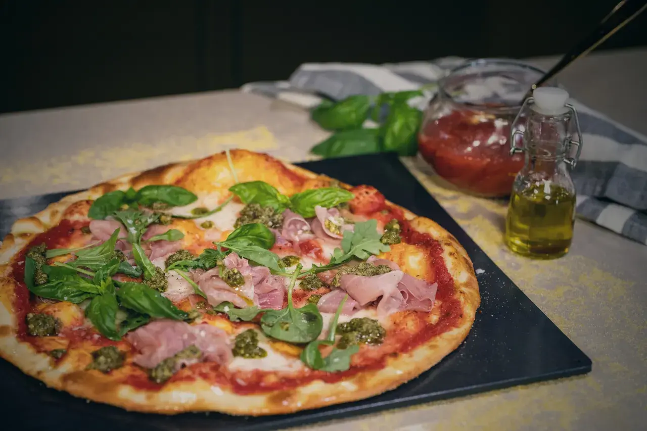 En läcker spenatpizza med prosciutto, basilika och pesto. Bredvid står en burk tomatsås och en flaska olivolja.