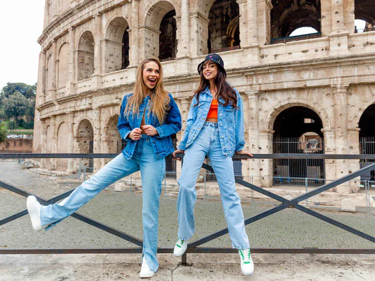 Dwie kobiety w strojach w kolorze denim pozują przed Koloseum. Jedna ma rozpuszczone włosy, druga nosi kapelusz.