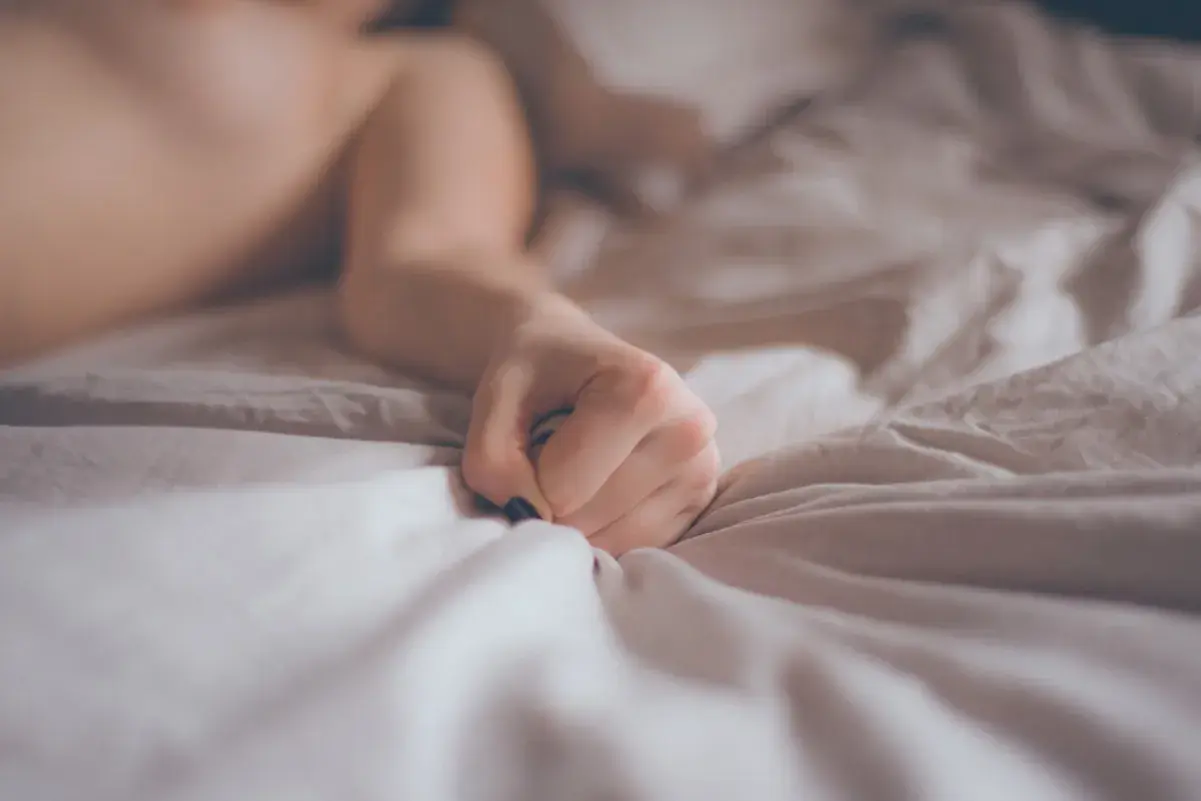 A person's hand clutches crumpled white sheets on a bed.