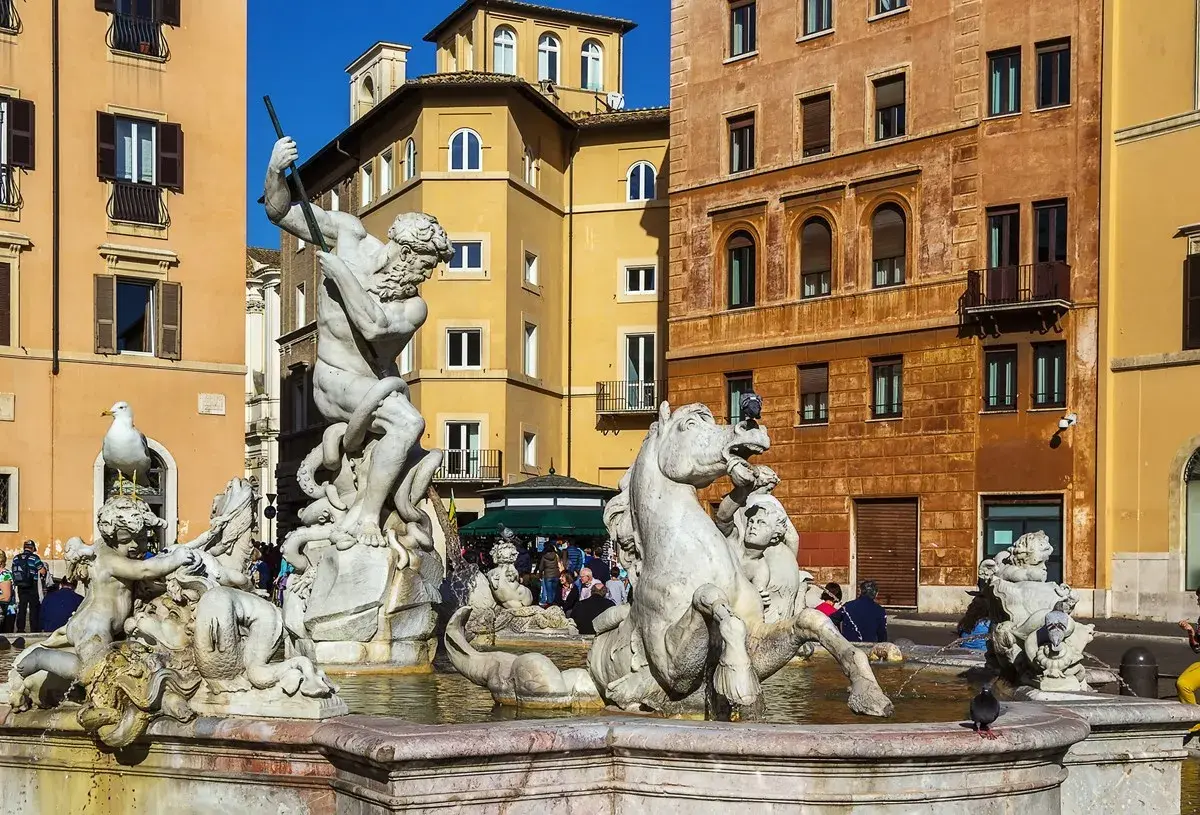 La Fuente de Neptuno en Roma, con su imponente estatua de mármol luchando contra un pulpo, rodeada de caballos marinos y figuras mitológicas, bajo el sol y edificios coloridos.