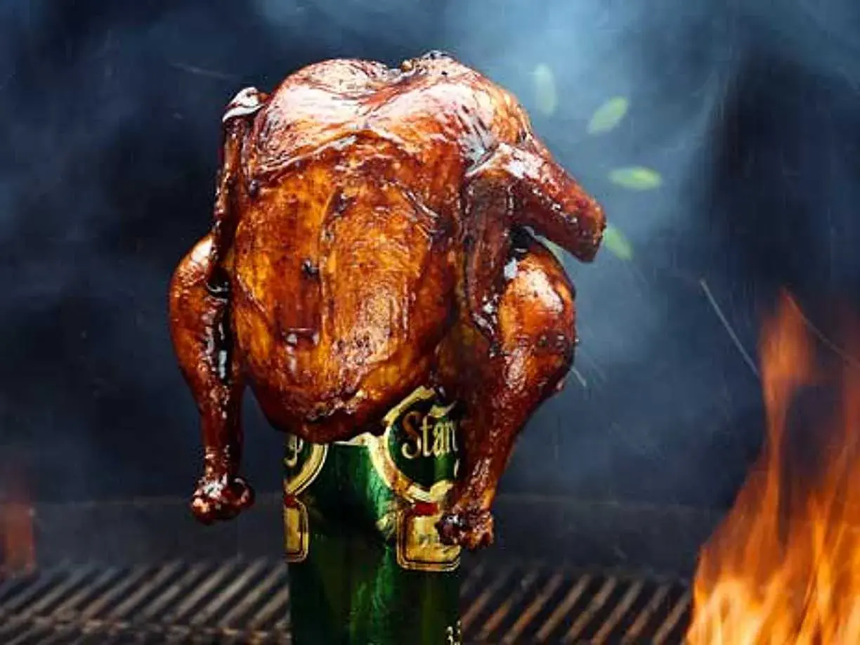 En saftig, gyllene hel kyckling tillagas stående på en ölburk, omgiven av rök och flammor. En klassisk beer can chicken.