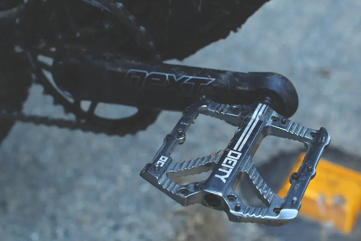 A close-up of a silver Deity pedal, ready for a Bladerunner-style descent. The crank arm reads "next SL.
