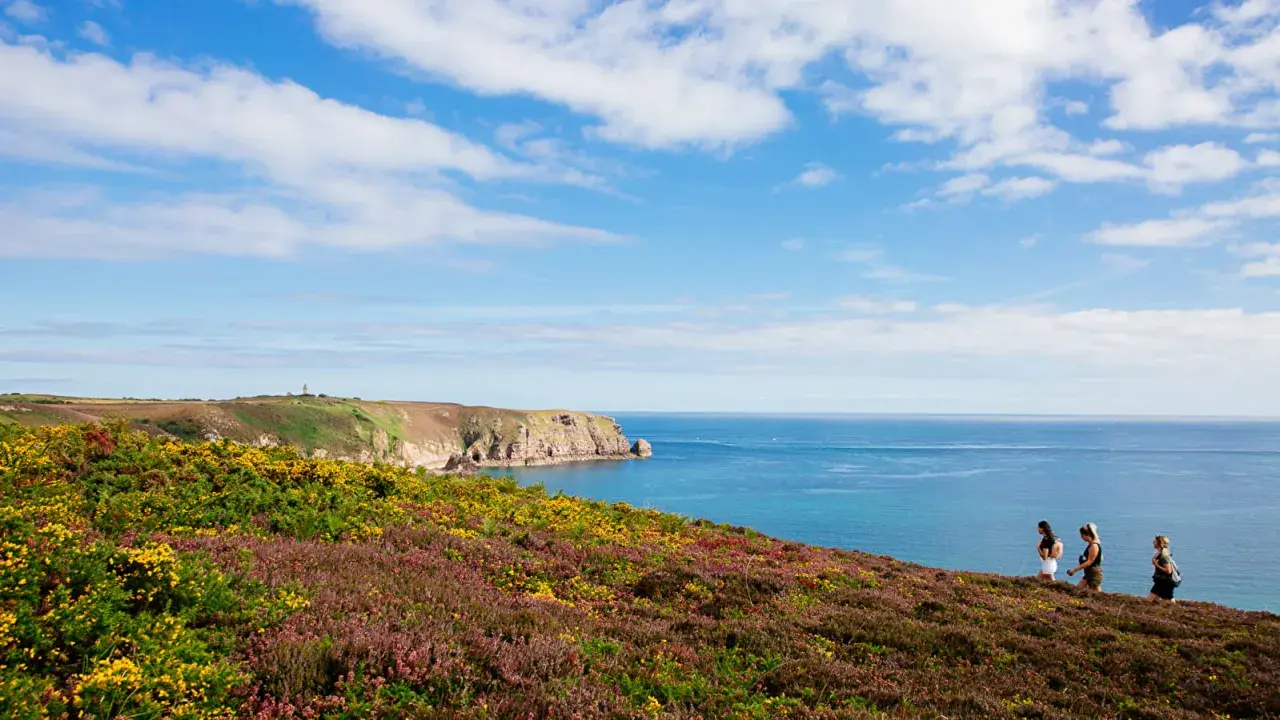 Trois femmes profitent d'un trek en Bretagne, longeant une côte rocheuse et fleurie, sous un ciel bleu parsemé de nuages.