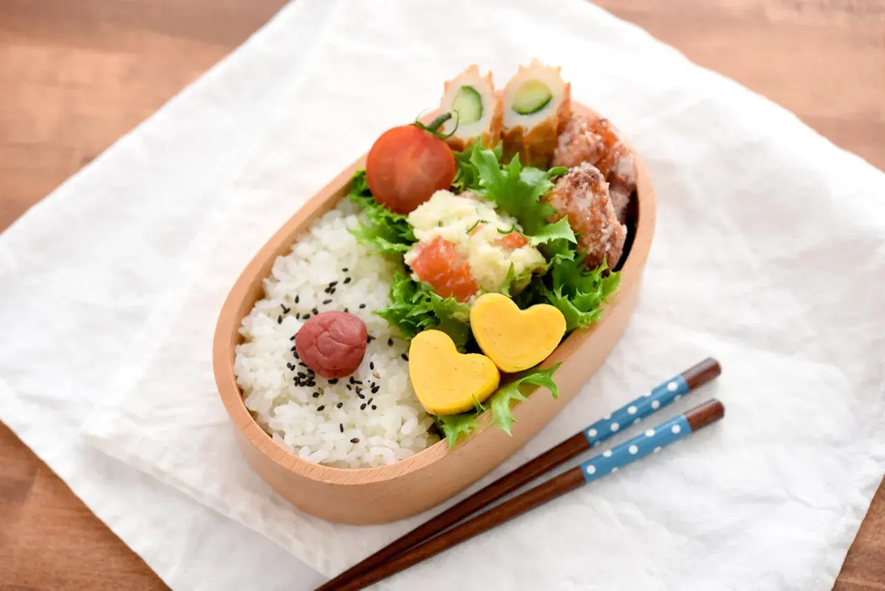 A delightful tamagoyaki bento box filled with rice, a pickled plum, potato salad, fried chicken, and cucumber rolls. Two heart-shaped tamagoyaki add a sweet touch.