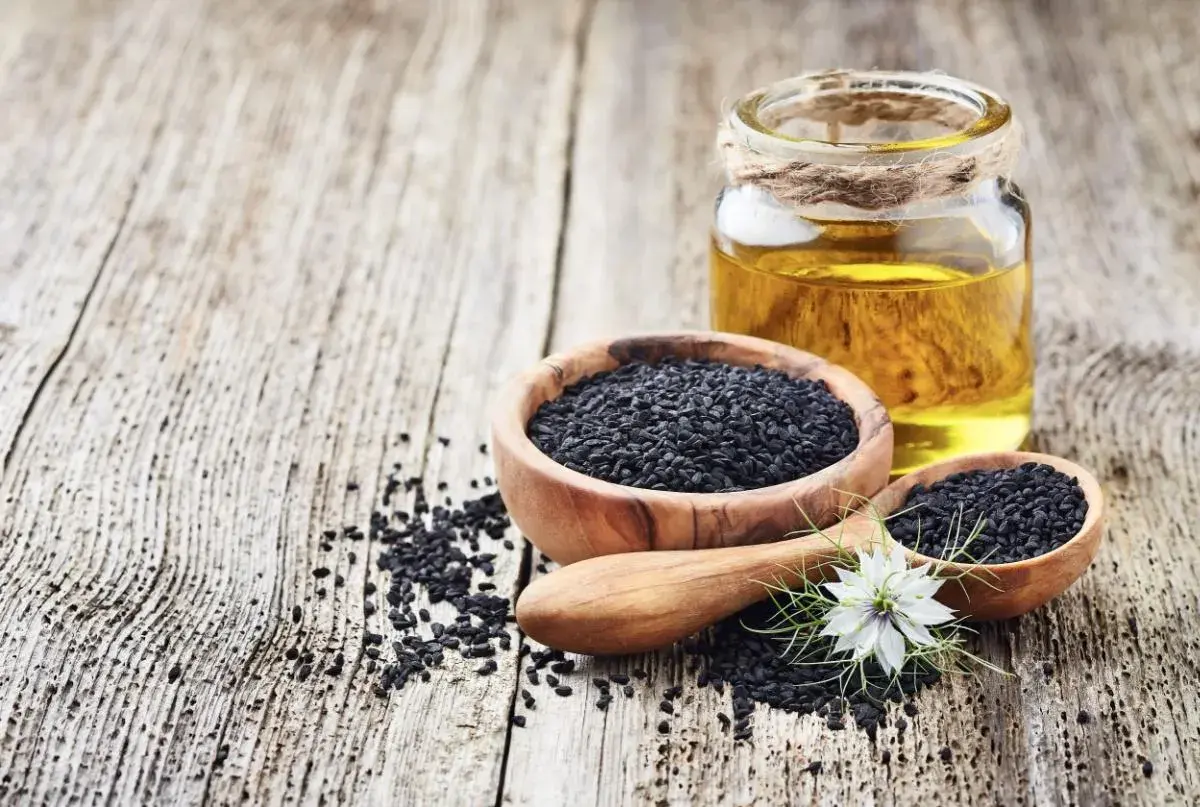 Semi neri in ciotole di legno e un flacone di olio di nigella, con un fiore bianco.