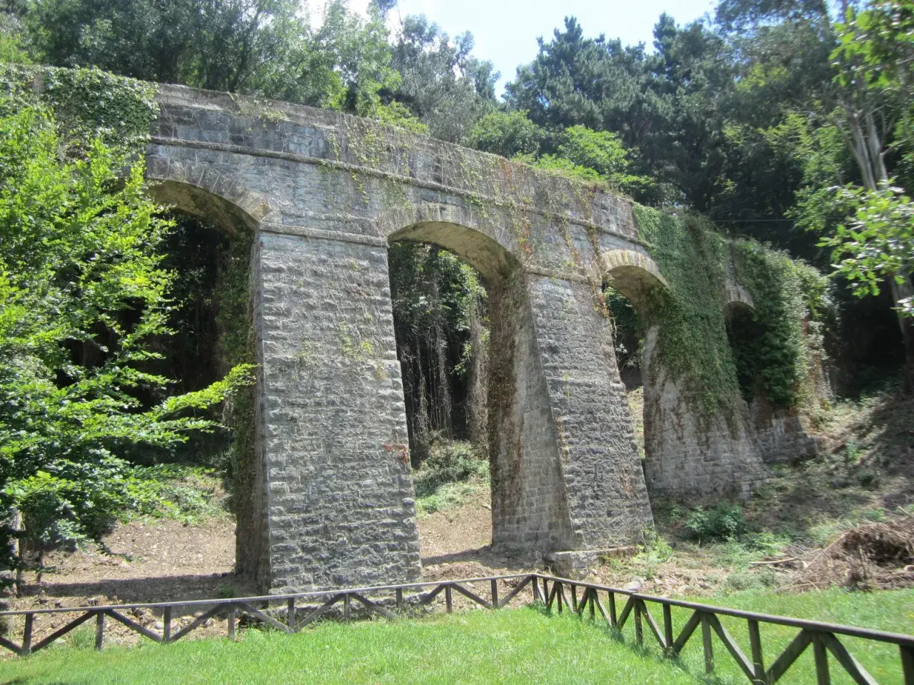 Antiguo acueducto de piedra cubierto de vegetación, con arcos y pilares robustos. Un camino verde pasa bajo su sombra.