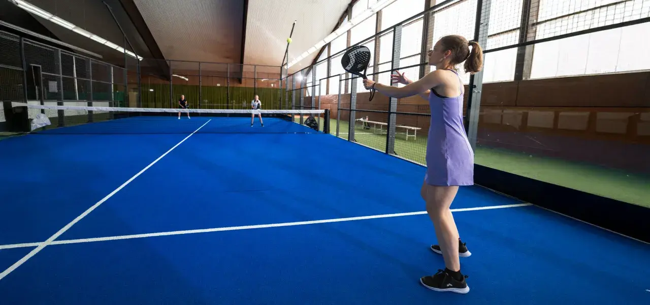 Ragazza in abito viola gioca a padel, con racchetta in mano, su campo blu. Altri giocatori in lontananza.