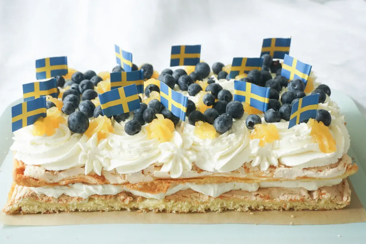 En stor brittatårta i långpanna, dekorerad med grädde, blåbär, apelsinbitar och små svenska flaggor. Perfekt för nationaldagsfirandet!