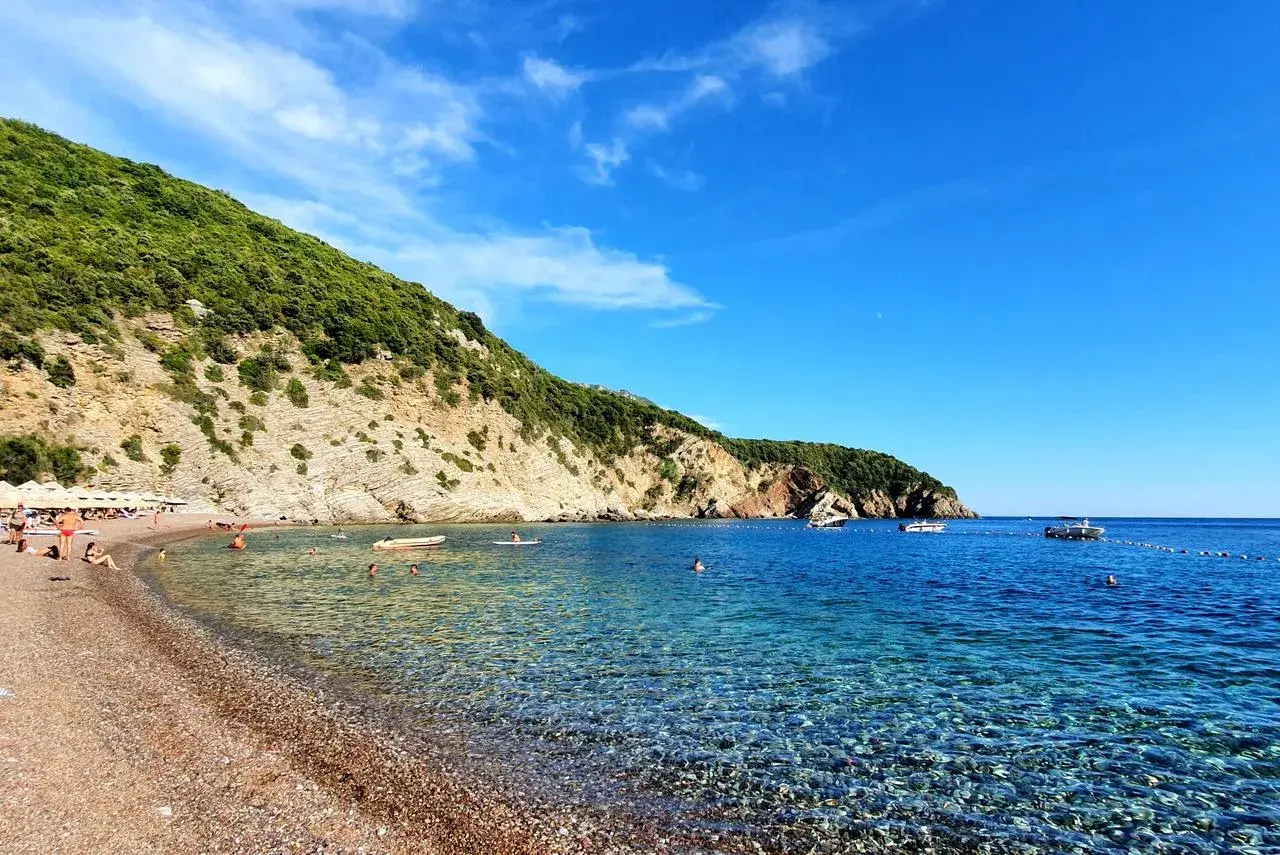 Kamienista plaża z turkusową wodą, zielonymi wzgórzami i kilkoma łodziami na morzu.